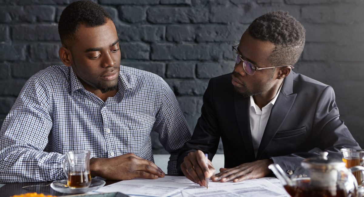 CEO in elegant suit and glasses pointing pen at papers on table in front of him, asking his financial expert to explain figures in report, staring at him with interrogative look. People and business