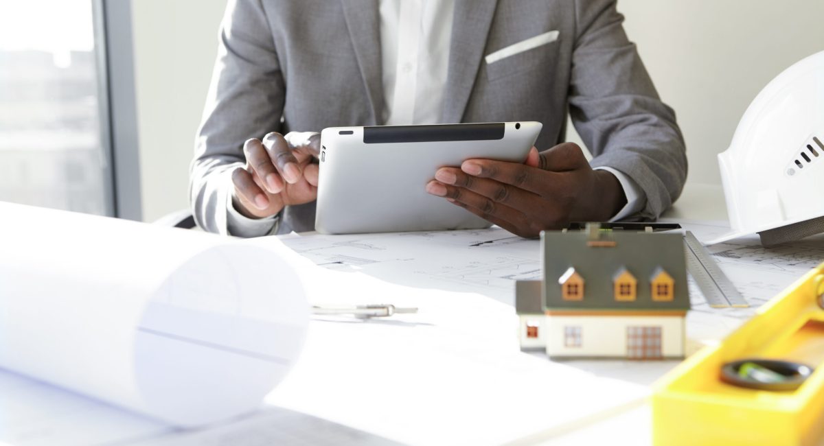 Selective focus. Design and architecture. Cropped shot of African architect holding digital tablet, developing new real estate project, sitting at desk with architectural tools and scale model house